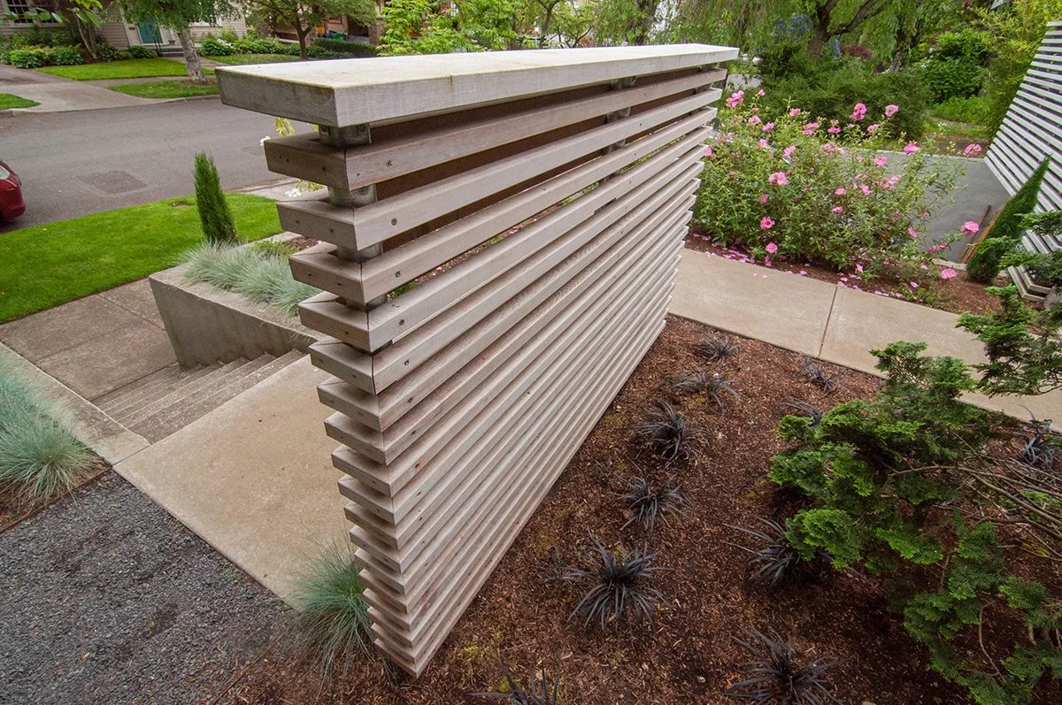 A modern wooden privacy screen on a front porch, with steps leading down to a landscaped yard with plants and pink flowers.