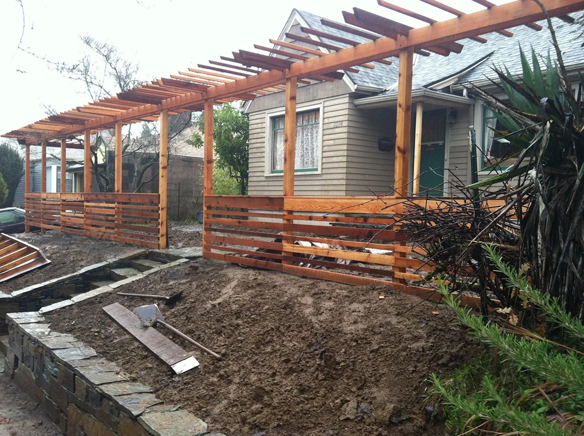 A house with a new wooden deck under construction, featuring wooden railing and pergola framework, with construction tools and dirt in the foreground.