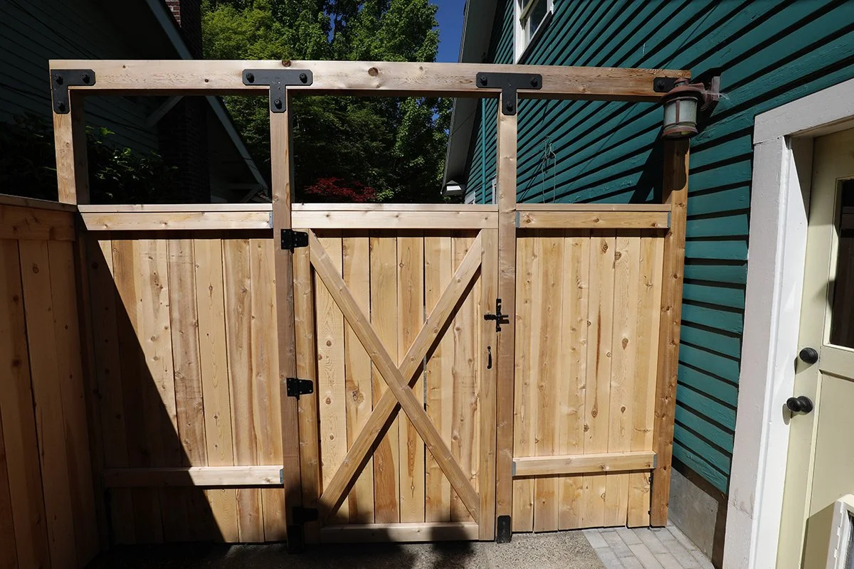 A newly constructed wooden gate and fence, installed between two houses, with a latch and hinges on the gate, and a lantern attached to the upper right corner of the fence.
