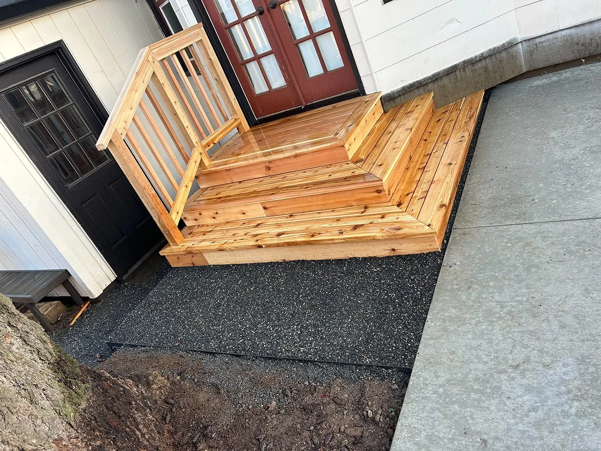 Wooden staircase with a railing leading up to a door on a house exterior, with a black door on the left and a red door in the background, adjacent to concrete and asphalt pavement.