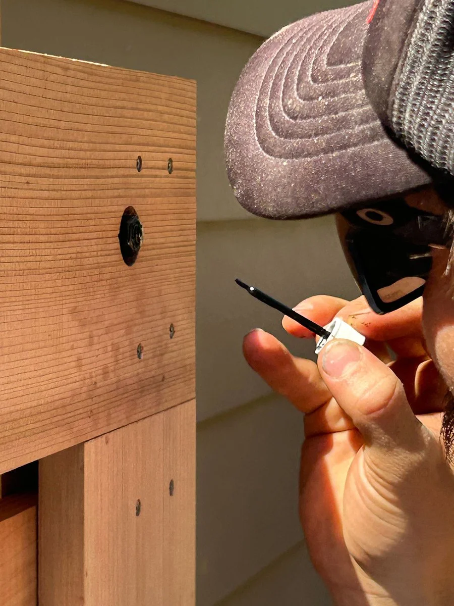 Person wearing a cap and sunglasses applying wood glue to a screw in a wooden furniture project.