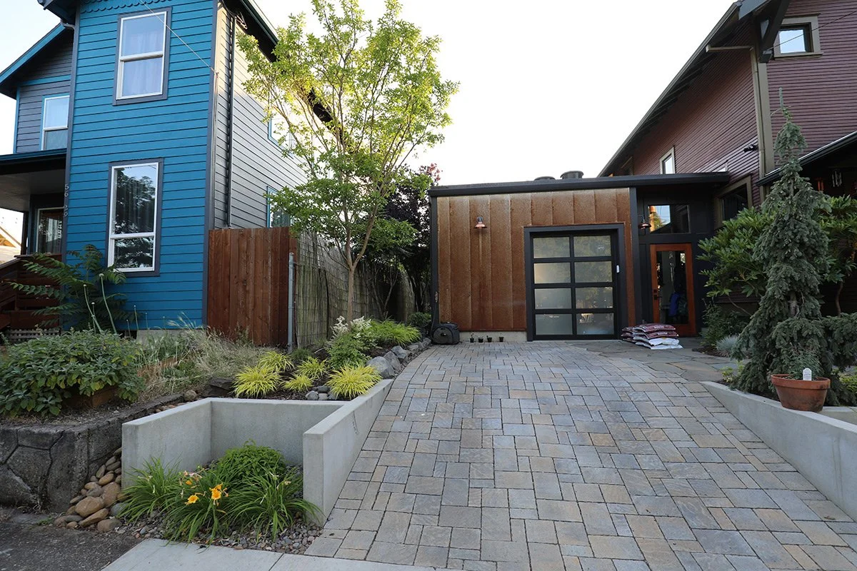 A modern residential backyard with a paved driveway leading to a garage door, surrounded by landscaped garden beds with various plants and trees, and two multi-story houses with colorful siding.