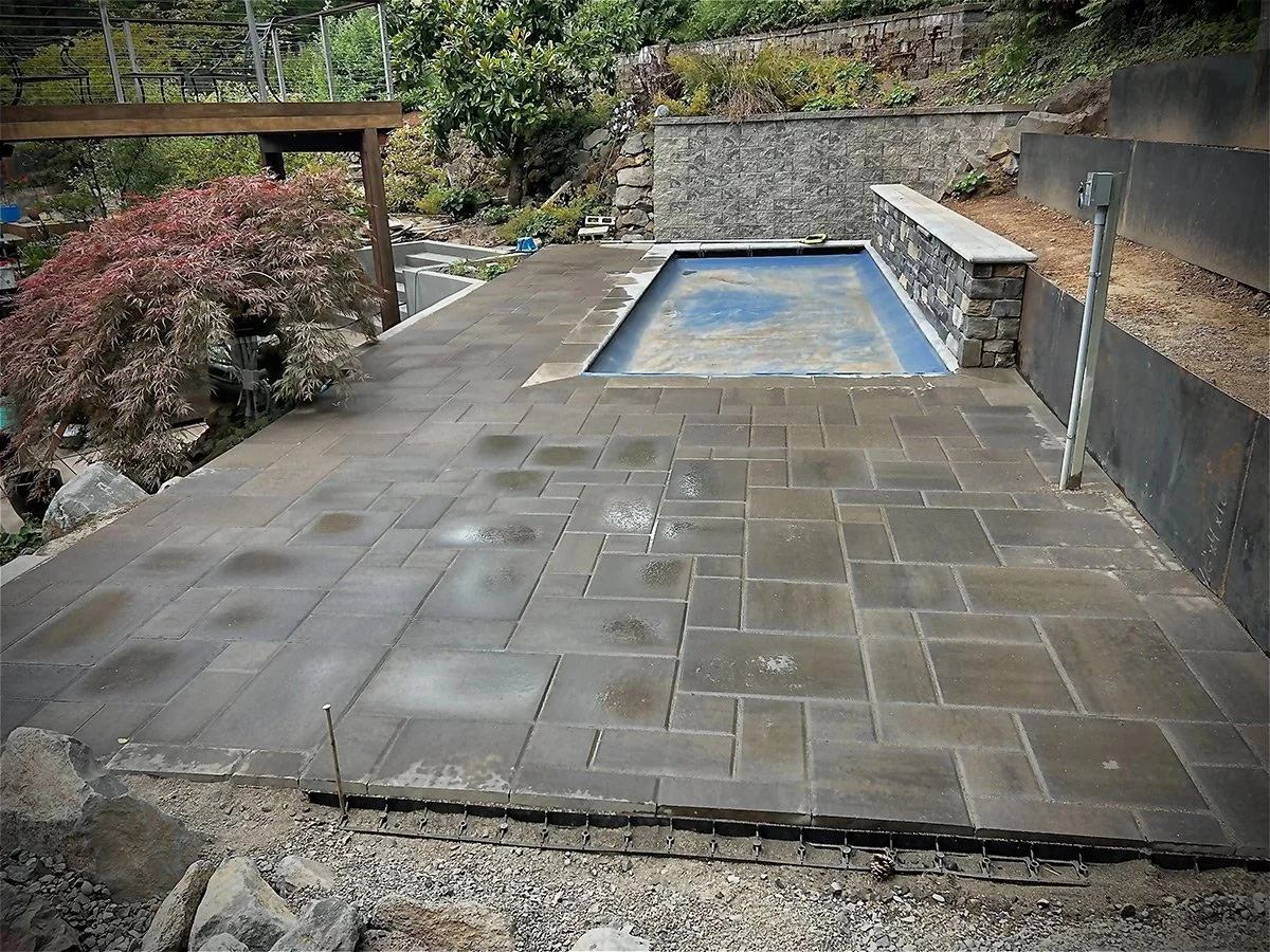 A newly paved patio with a small rectangular pool and a stone wall in a backyard garden.