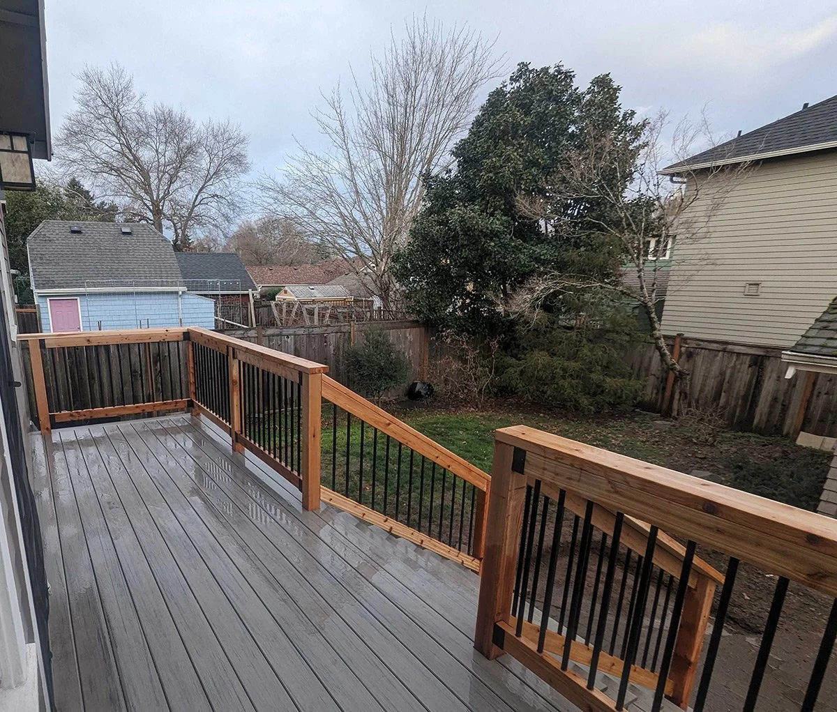 View from a new wooden deck with black metal railing, overlooking a backyard with trees and neighboring houses, some with gabled roofs and siding. The sky is overcast.