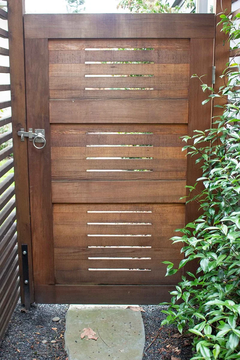 Wooden gate with horizontal slats, secured with a steel latch, surrounded by green bushes and a stone step at the bottom.