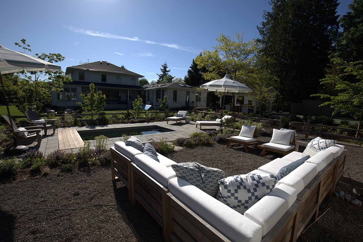 Backyard with outdoor seating area, lounge chairs, umbrellas, and garden, with a swimming pool and house in the background.