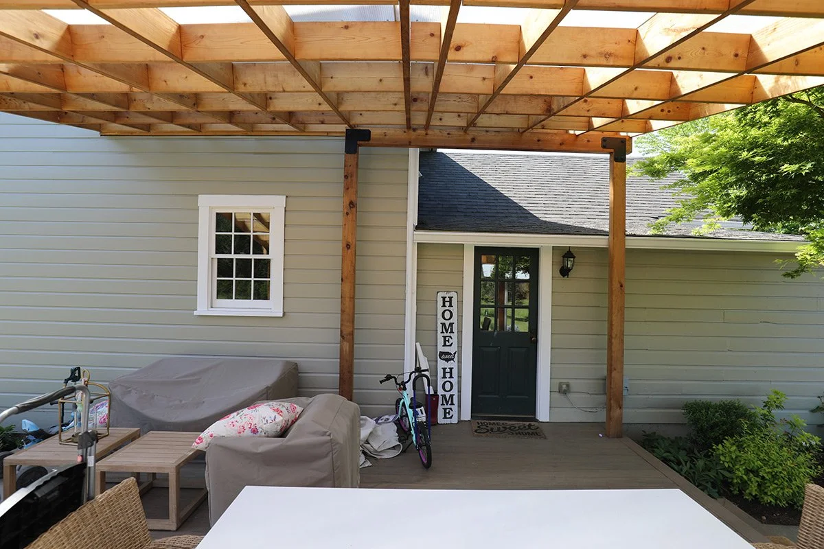 Covered porch area with a new wooden pergola, a green front door with glass panes, a window, outdoor furniture, children's bike, and decorative signs that say "Home".