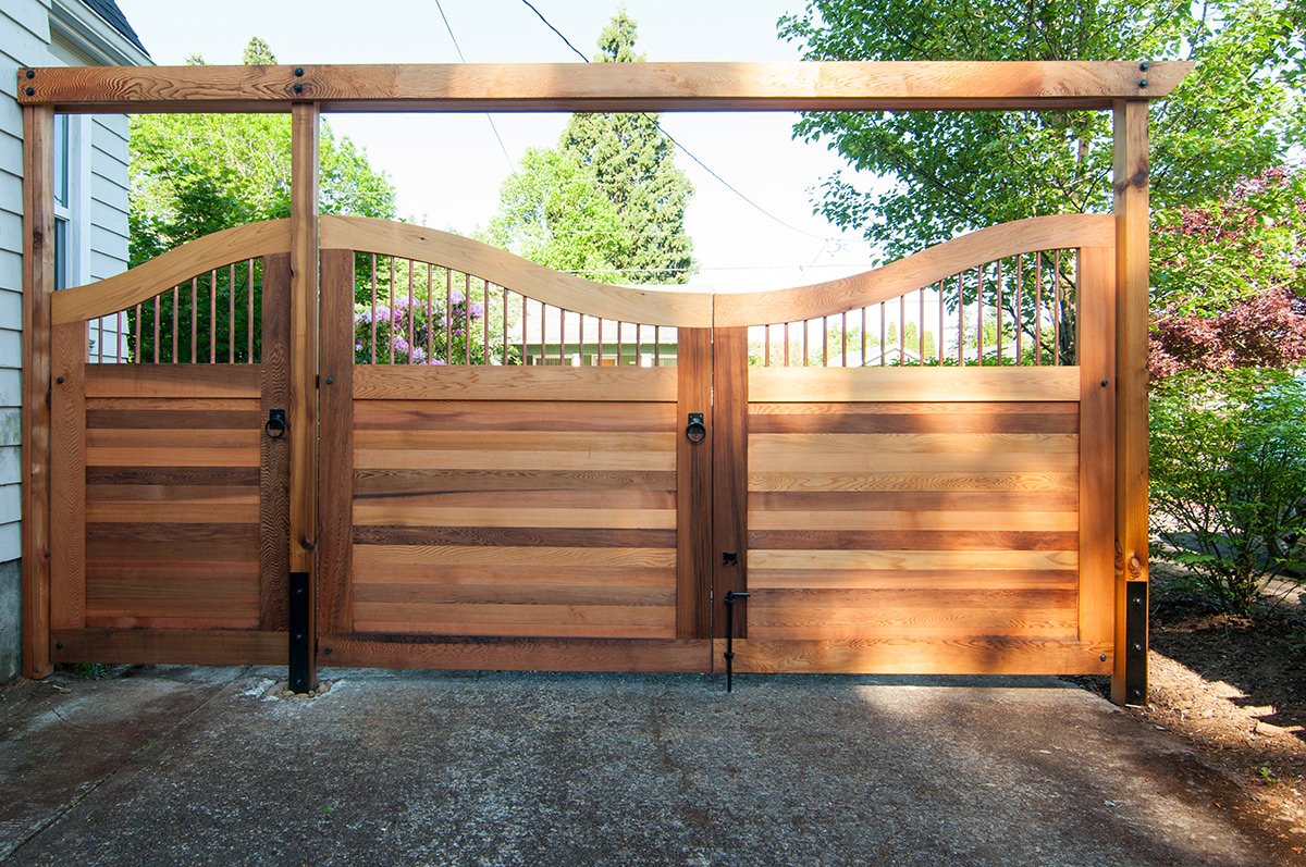 A wooden gate with decorative curved top panels, installed on a concrete driveway next to a house with light blue siding, surrounded by green trees and bushes.