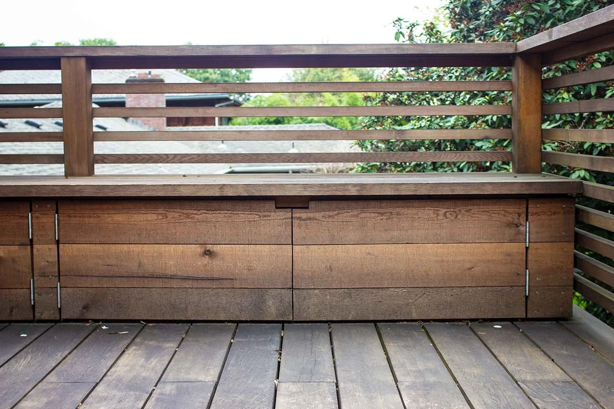 A wooden outdoor bench with a storage compartment, located on a deck with wooden planks, and surrounded by a wooden railing.