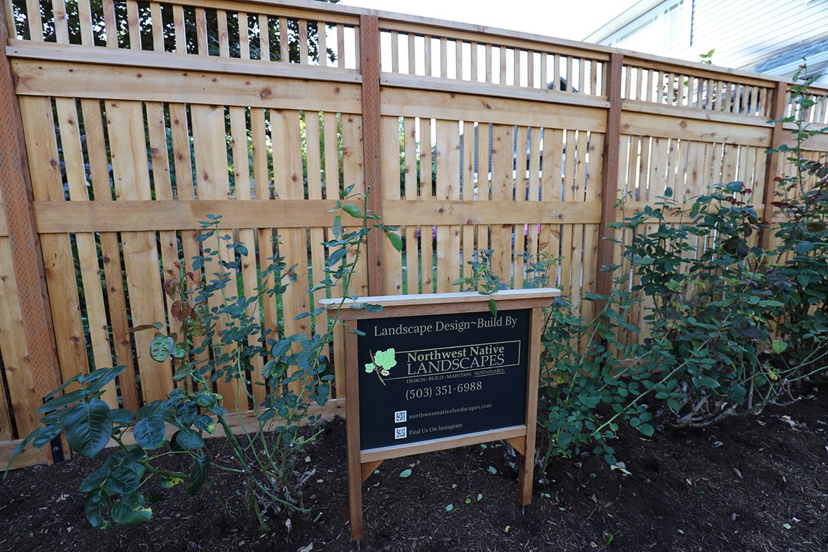 A newly built wooden privacy fence with horizontal and vertical slats surrounds garden beds with green plants and roses. A sign in front of the fence reads 'Landscape Design Build By Northwest Native Landscapes' along with contact information and soc