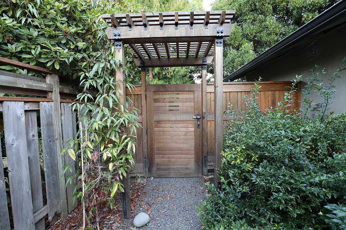 Wooden gate with a pergola on top, surrounded by green bushes and trees, with a small gravel pathway leading to it.