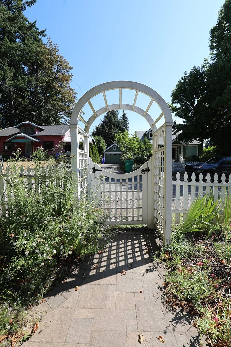 White garden gate with an arched top, cast shadows on brick pathway, surrounded by greenery and plants in a residential neighborhood under a clear blue sky.