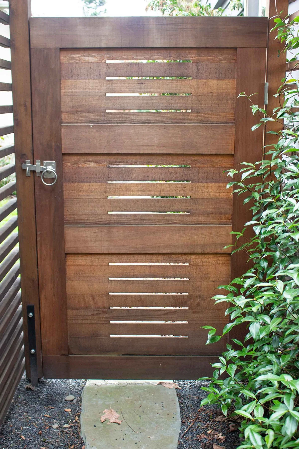 A wooden gate with horizontal slats, secured with a round lock, surrounded by greenery and gravel ground with a concrete stepping stone.