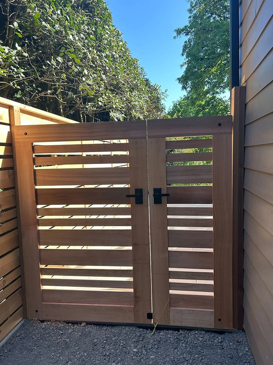 A newly built wooden backyard gate with slats and black hinges, set between wood and vinyl fences under a bright blue sky with trees in the background.