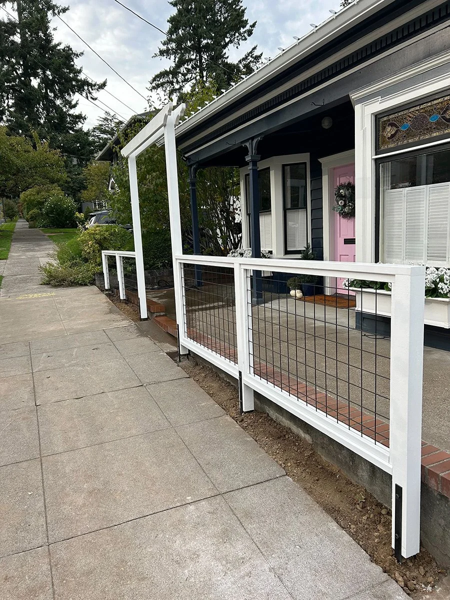 A newly installed white fence with wire mesh along a sidewalk in front of a house with a purple front door, porch, and decorative stained glass window.