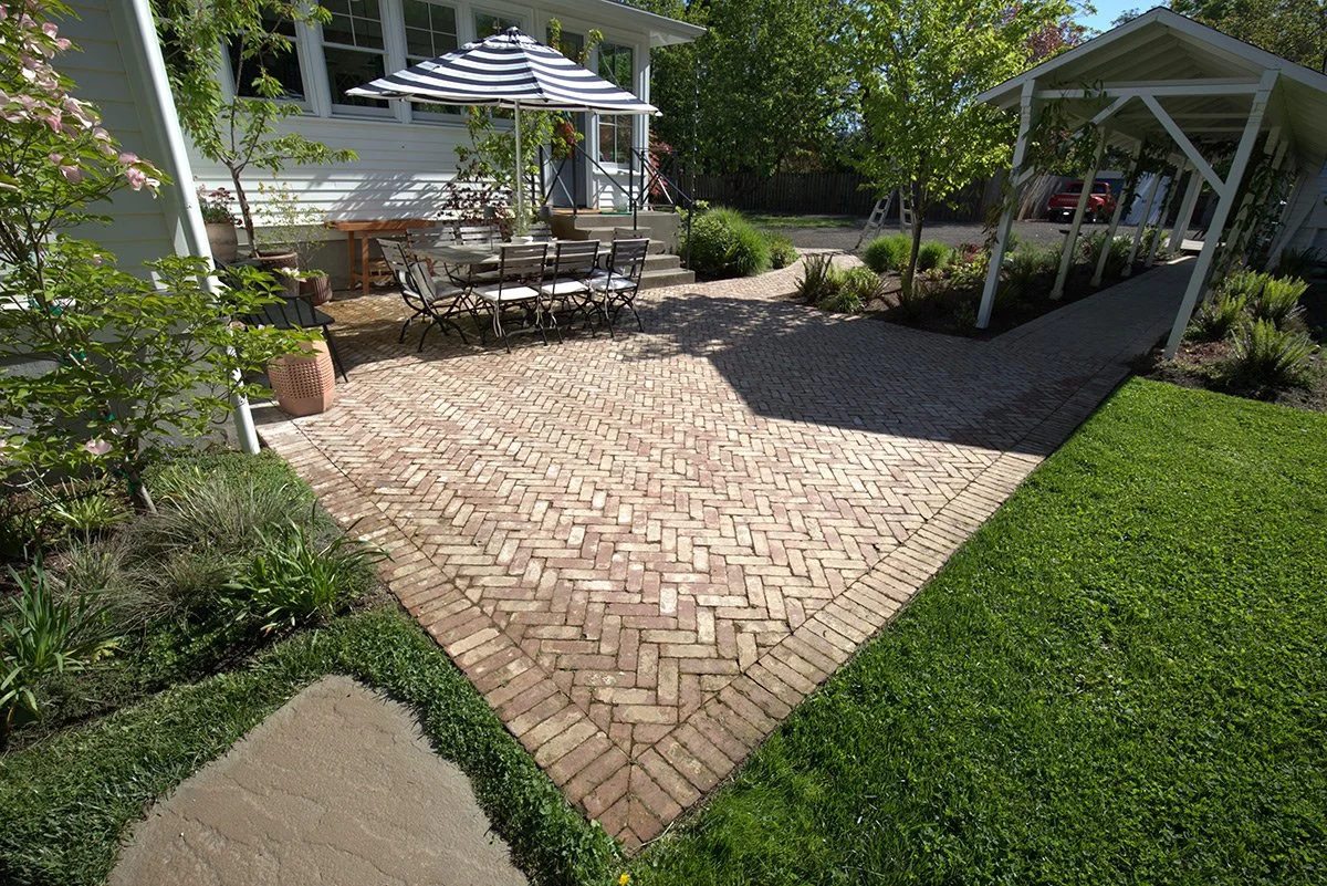 A brick patio with outdoor dining furniture, including a table, chairs, and a striped umbrella, in a garden backyard with trees, plants, a pathway, and a carport.