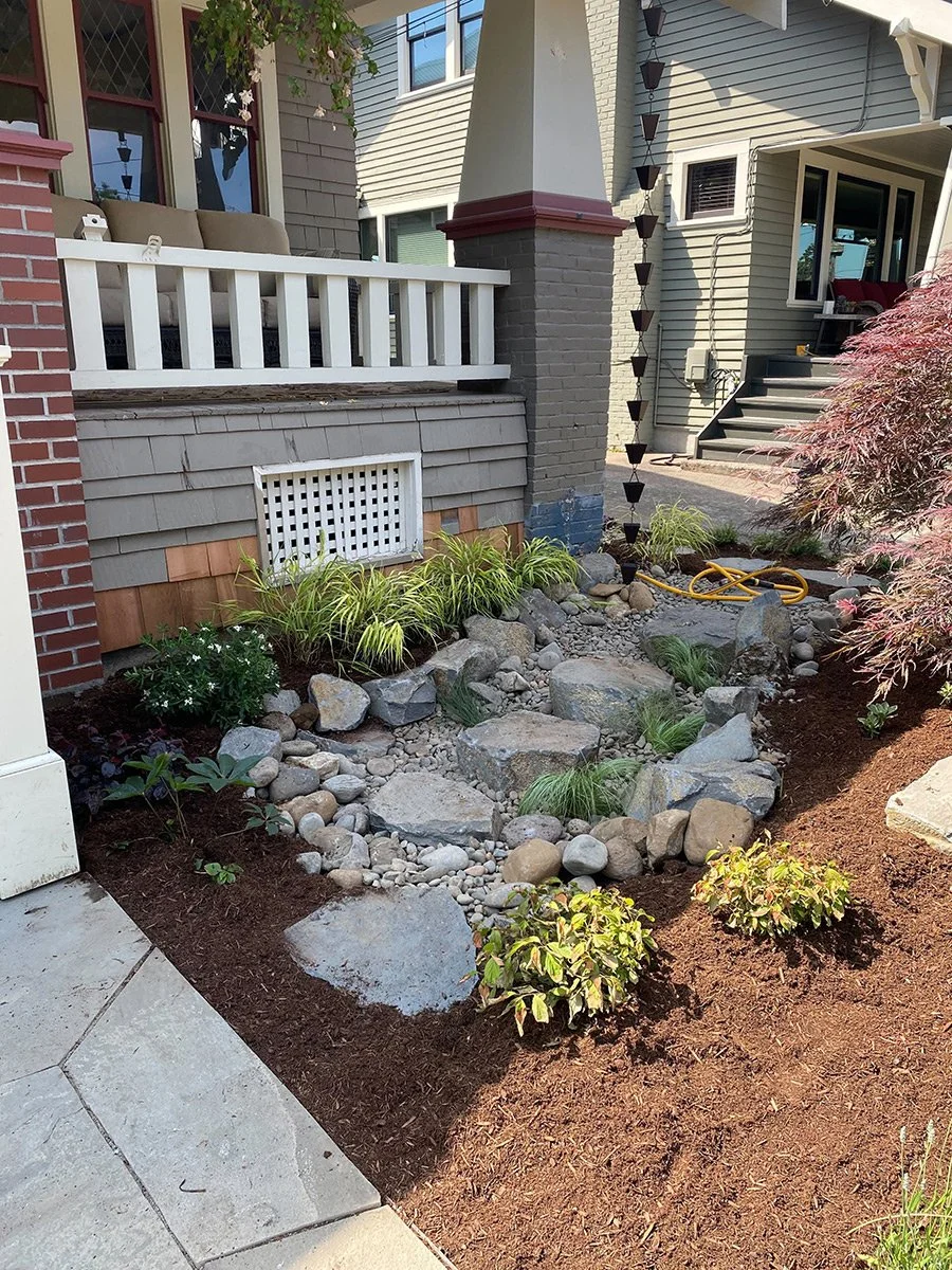 A landscaped garden area with rocks, plants, and mulch next to a house with stairs and a porch.