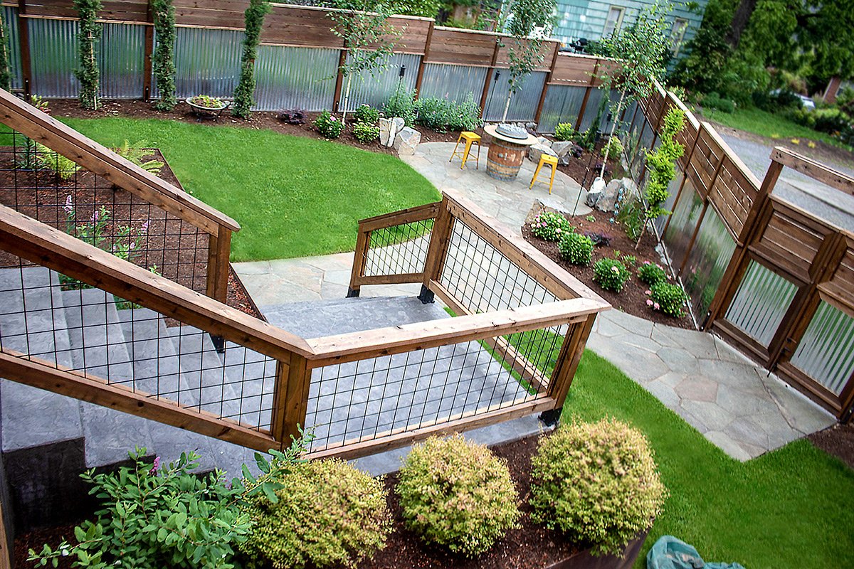 A backyard with a wooden deck and railing, green lawn, flower beds with shrubs and flowers, a stone patio area with yellow stools and a table, and a picket fence with trees along it.