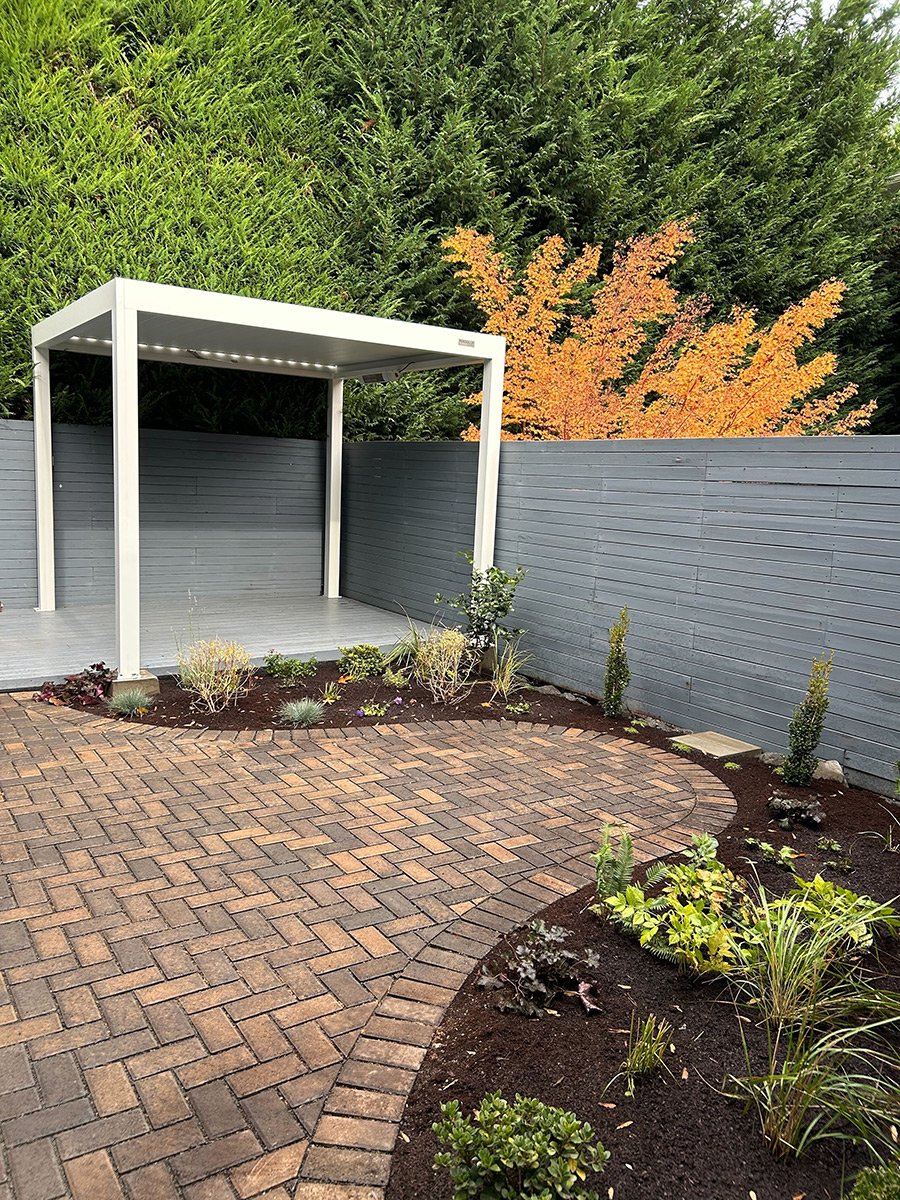 Small backyard garden with a paved brick patio, a gray fence, and a white pergola with built-in lighting, surrounded by various plants and bushes, with tall green trees and a fall-colored tree in the background.