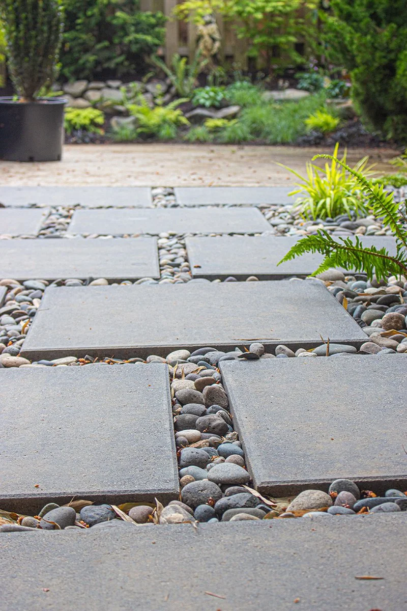 Stone pathway with large rectangular pavers and small round stones, surrounded by greenery and plants in a garden.