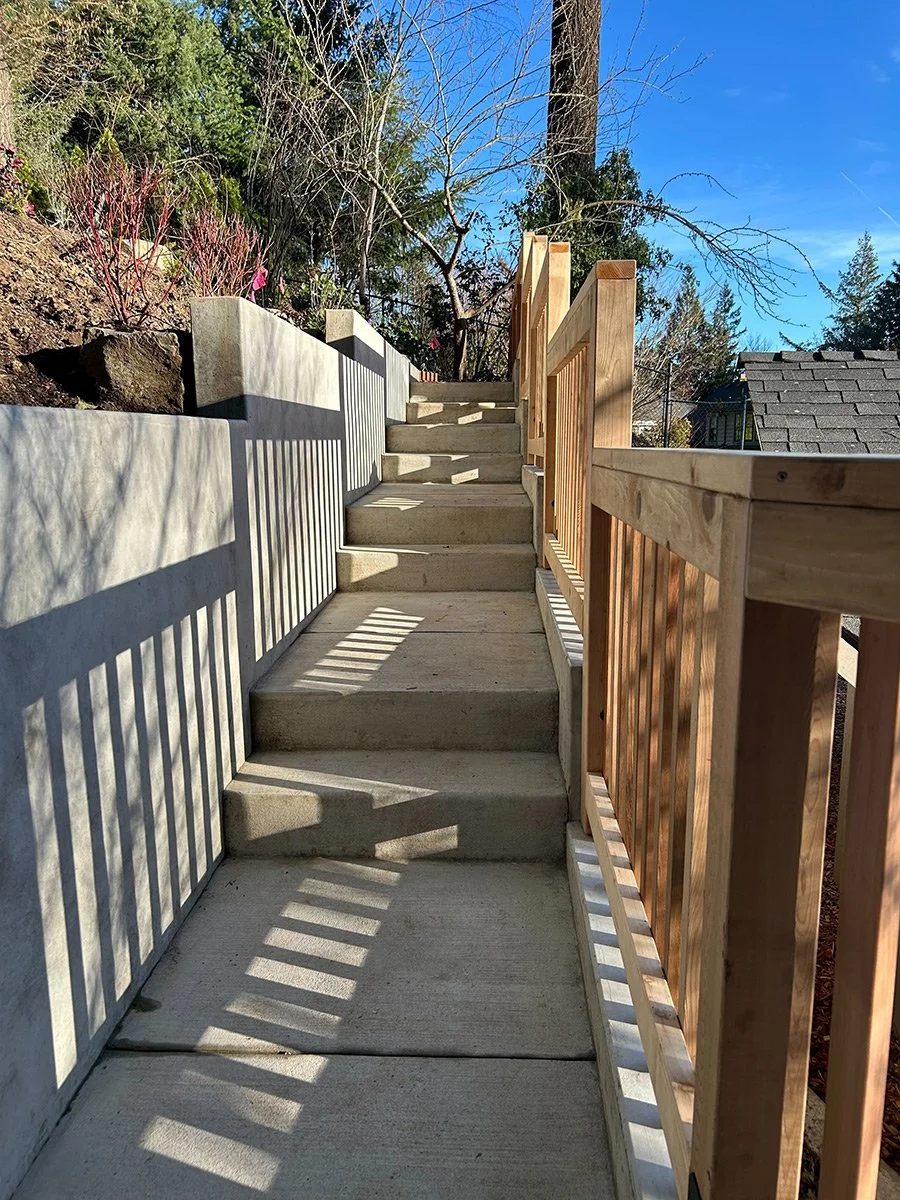 Concrete stairs leading upward with a planter on the left and a wooden railing on the right, shadows of the railing cast on the stairs, trees and a house roof visible in the background under a clear blue sky.