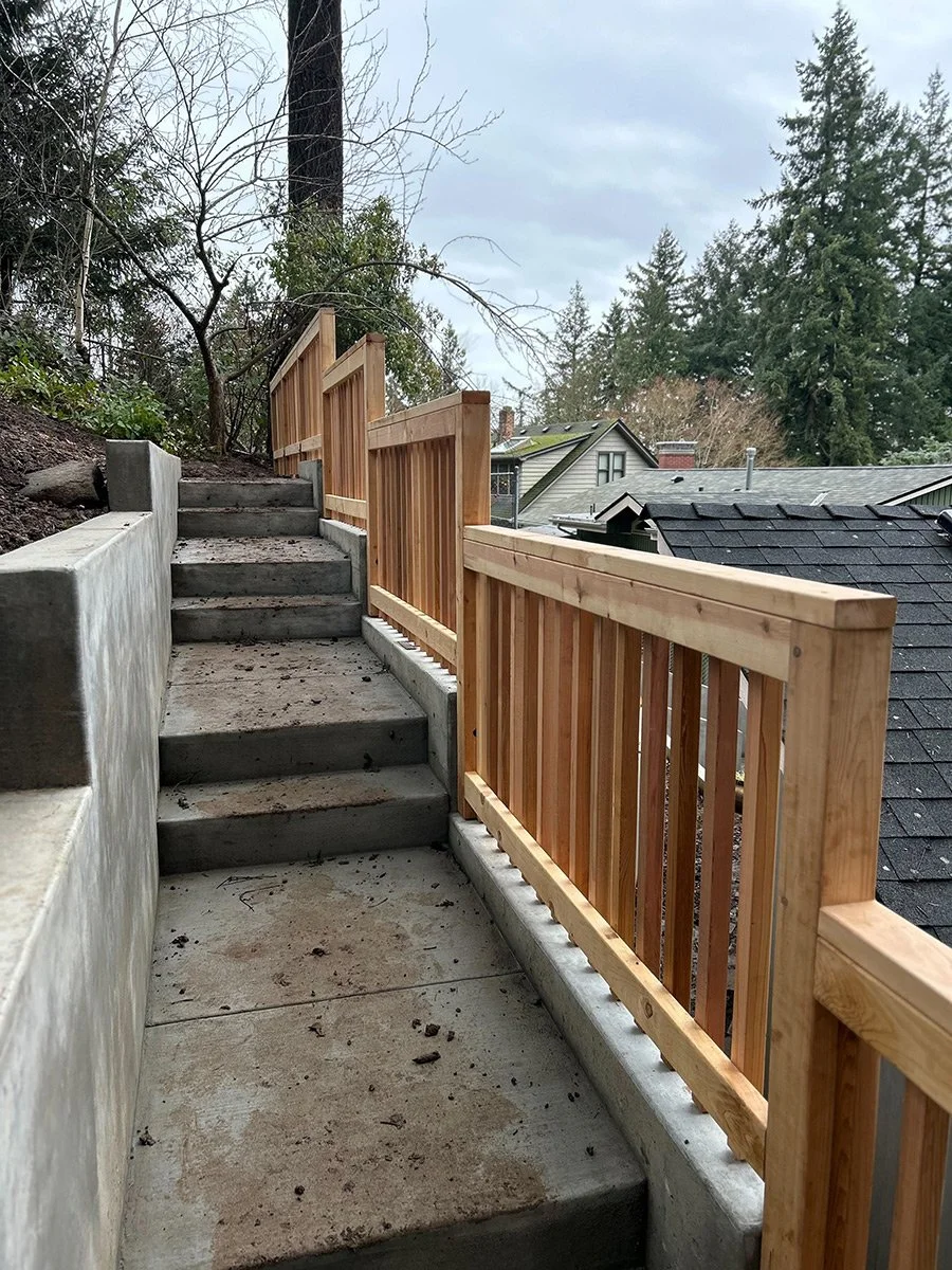 Concrete stairs with a wooden railing on one side, leading up a slope on a residential property with trees in the background.