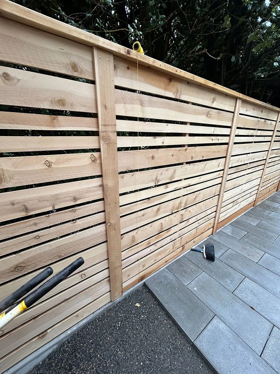 Wooden slat privacy fence under construction next to paved pathway, with tools lying on the ground nearby.