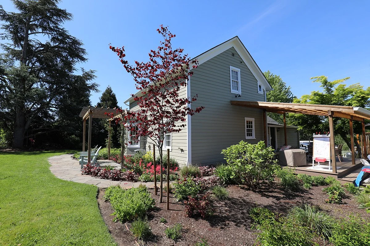 A two-story house with gray siding, surrounded by a landscaped yard with green grass, trees, and flowering plants. There is a wooden patio with outdoor furniture and a pergola on the porch.