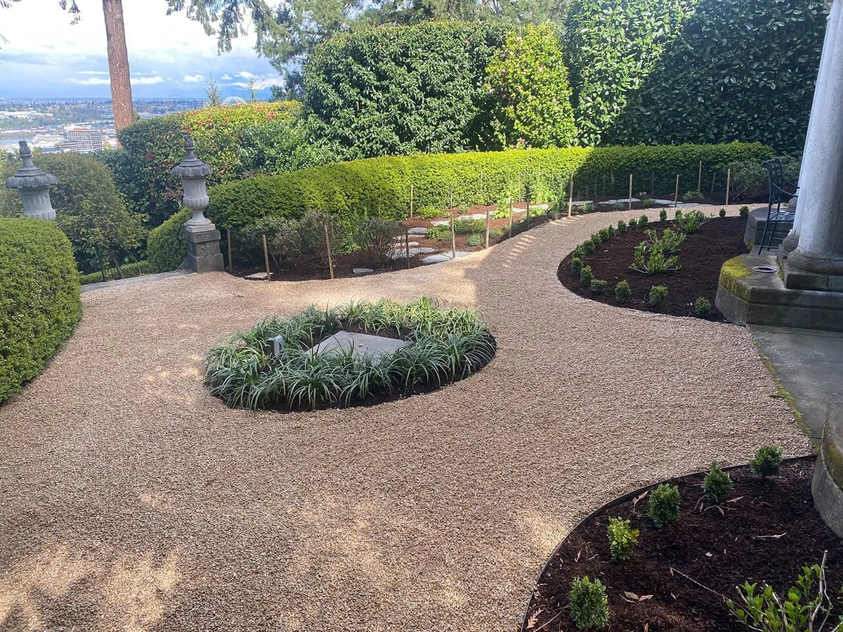 A landscaped garden walkway with trimmed bushes, small plants, and decorative stone lamps. Tall trees and cityscape visible in the background.