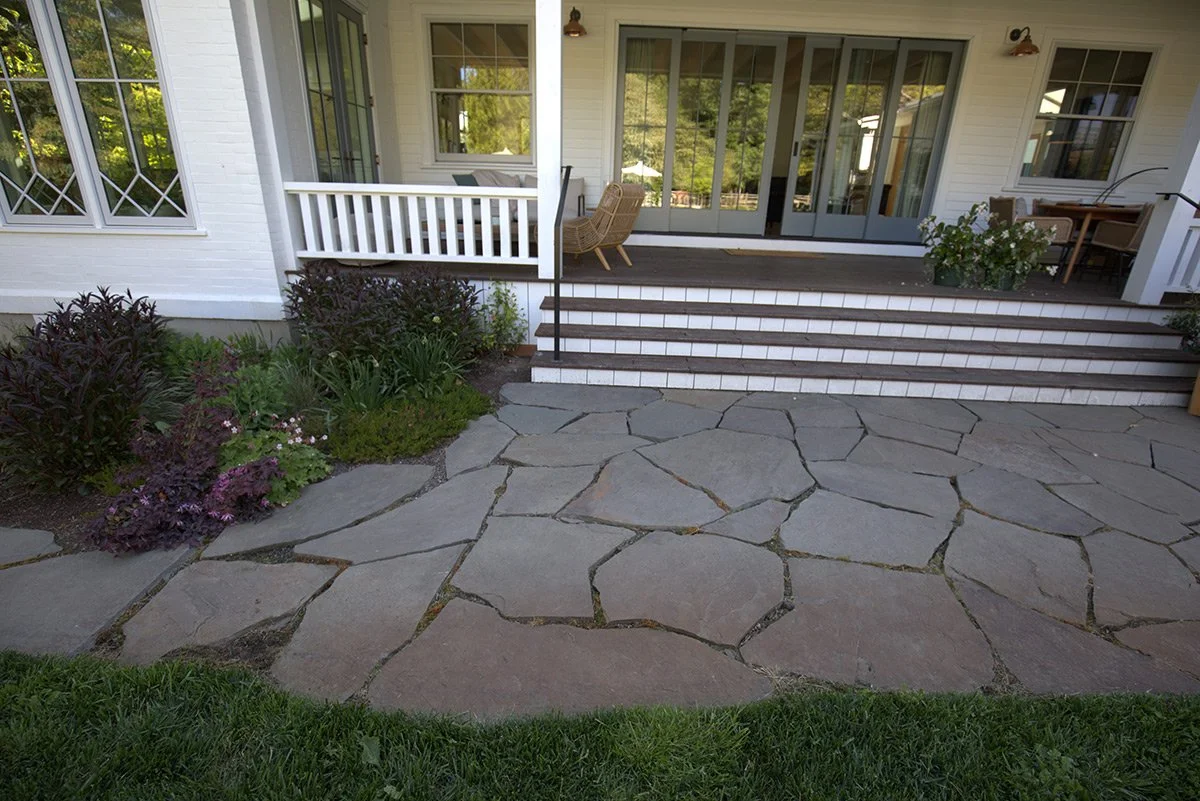 View of a house backyard with a stone patio, steps leading up to a screened porch, and a garden with bushes and plants.