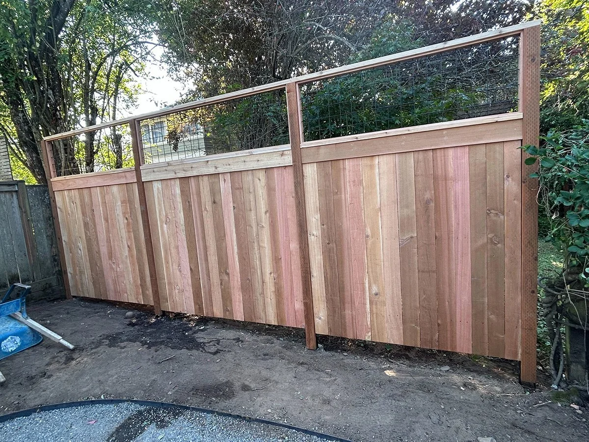 A wooden fence with a combination of horizontal and vertical slats, topped with wire mesh, in a backyard with trees in the background.
