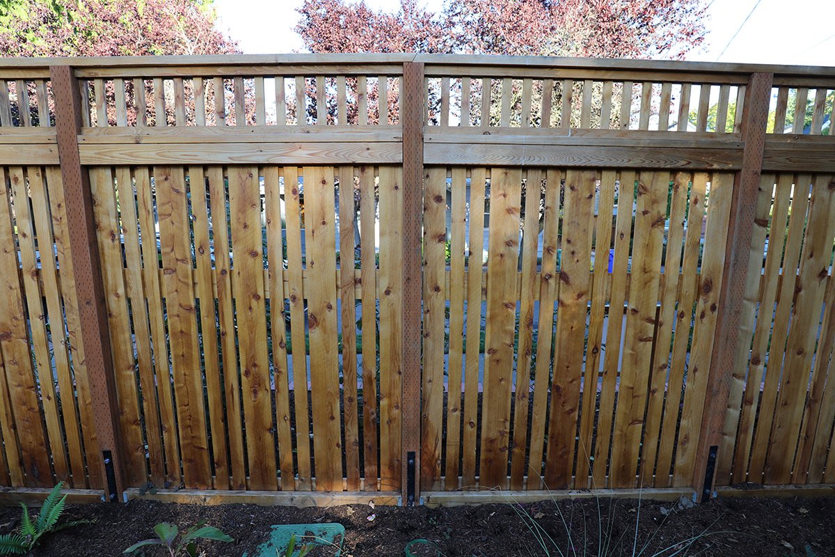 New wooden fence with vertical slats and lattice top, installed in a backyard, with some greenery and trees visible in the background.