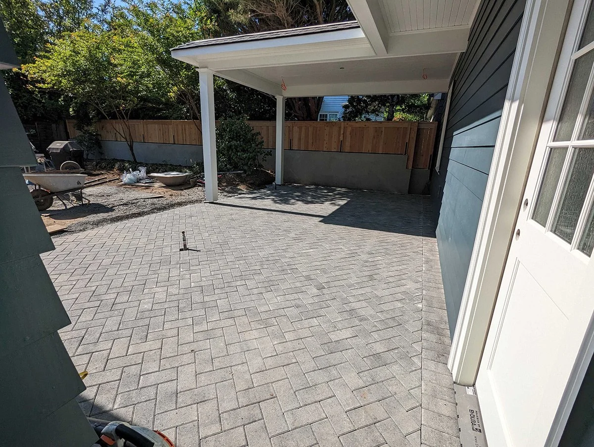 Backyard patio with newly installed brick pavers, part of a house with a covered patio area, a white door, a wooden fence, and some trees and planting in the background.