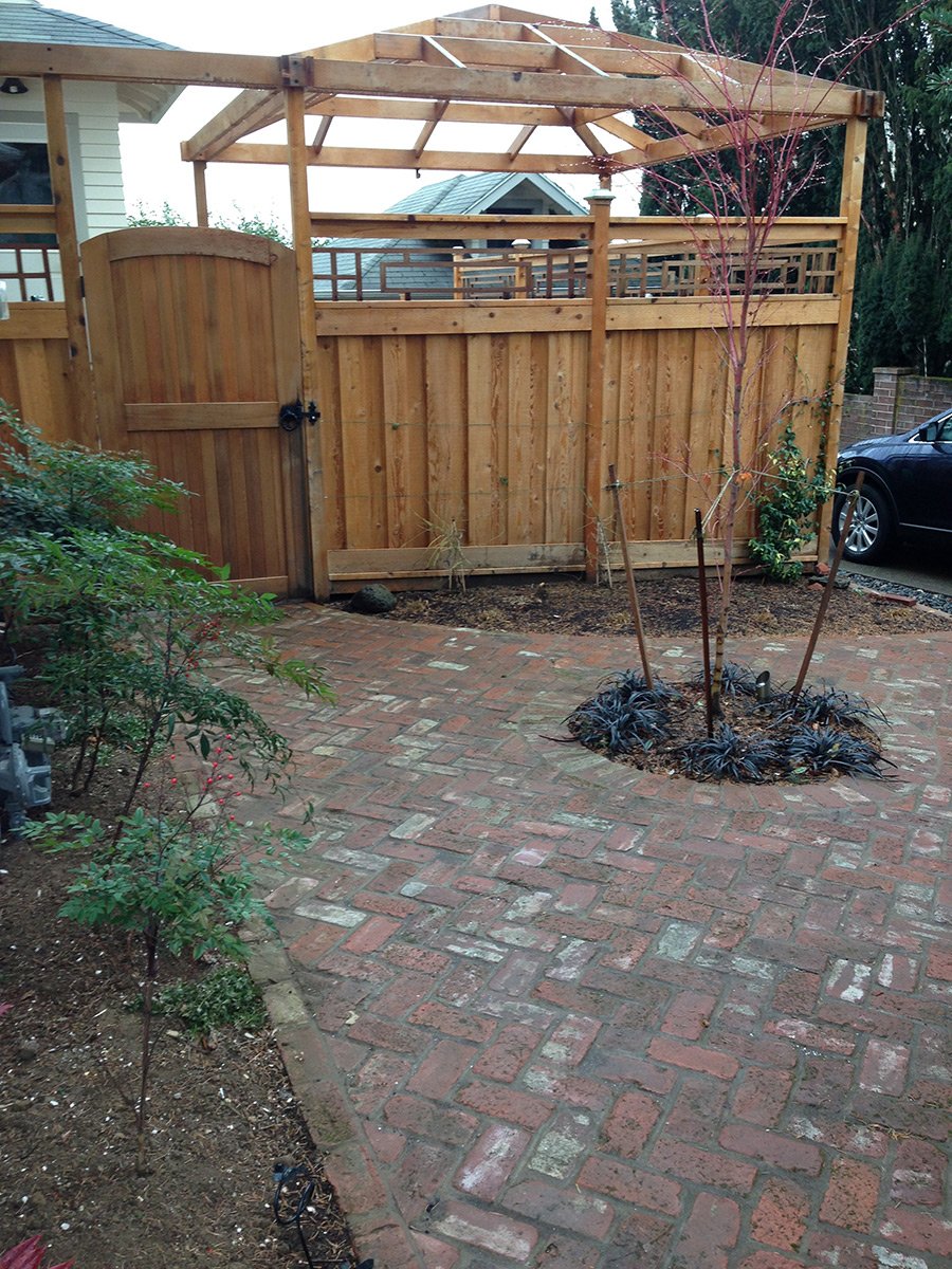 A backyard with a brick patio, a small tree with reddish branches, and a wooden fence with a gate. The fence has an upper lattice section, and the top framework for a future roof or cover is visible. There is a parked car to the right and some shrubs