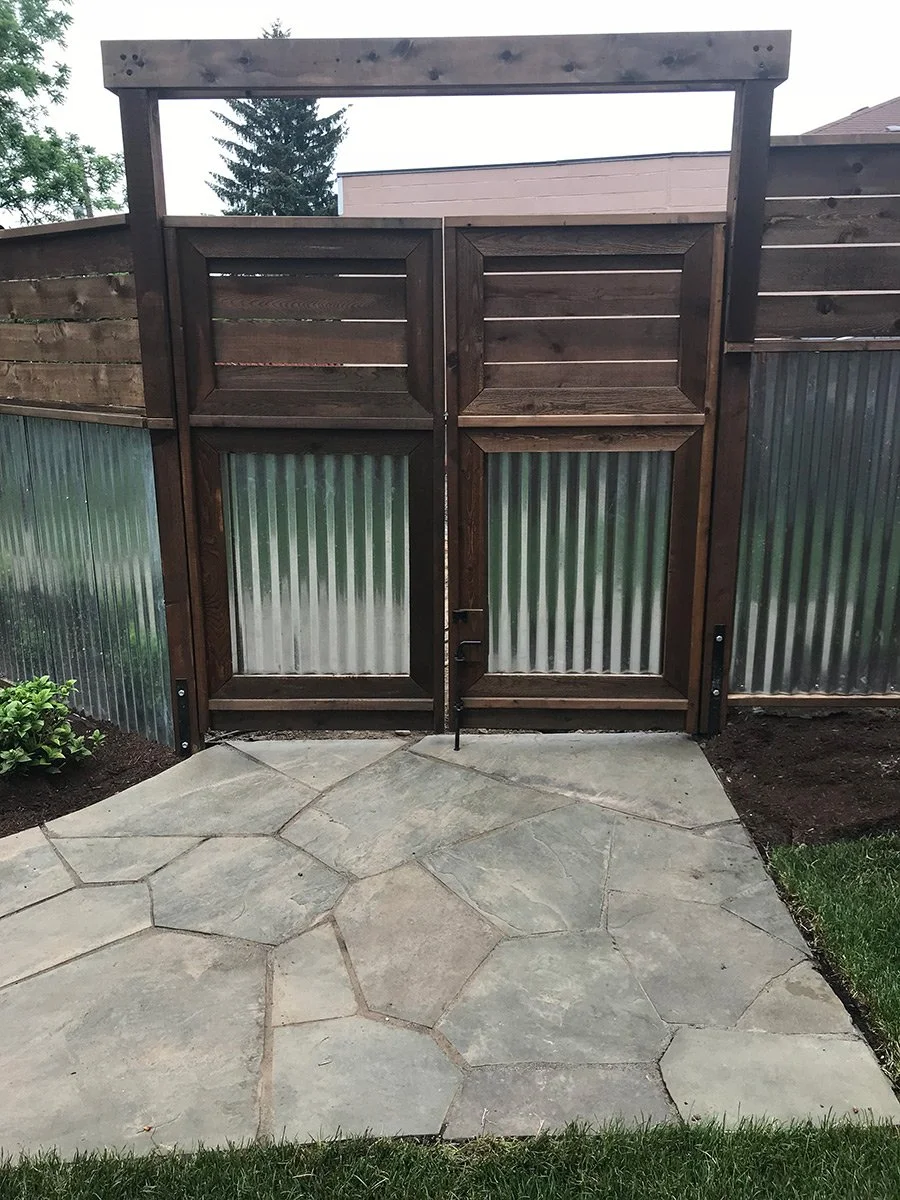 A small backyard area with a stone paved patio and a wooden gate with corrugated metal panels, surrounded by a wooden fence and some greenery.