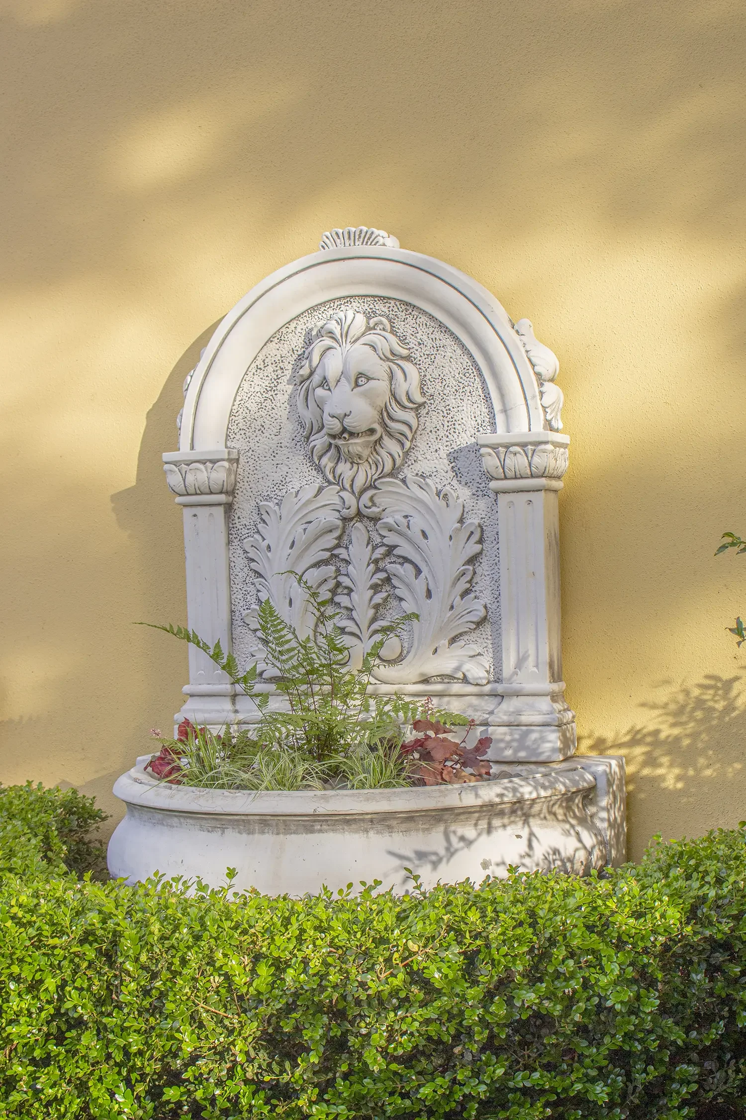 Decorative white stone fountain with a lion's head relief, set against a yellow wall, surrounded by green bushes and plants.