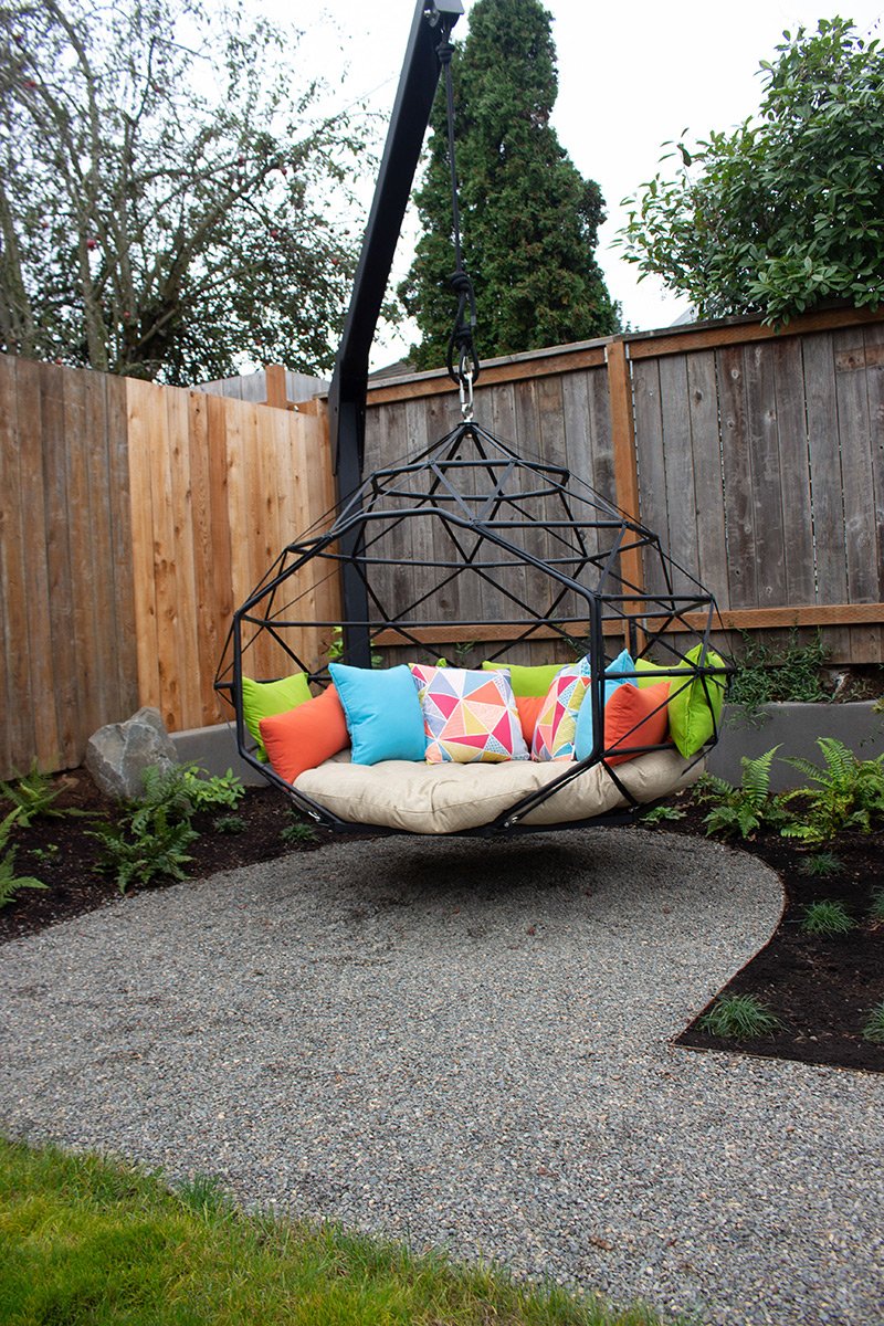 Outdoor hanging swing with colorful pillows on a cushioned seat, suspended from a black support, in a garden area with gravel pathway, green grass, plants, and a wooden fence.