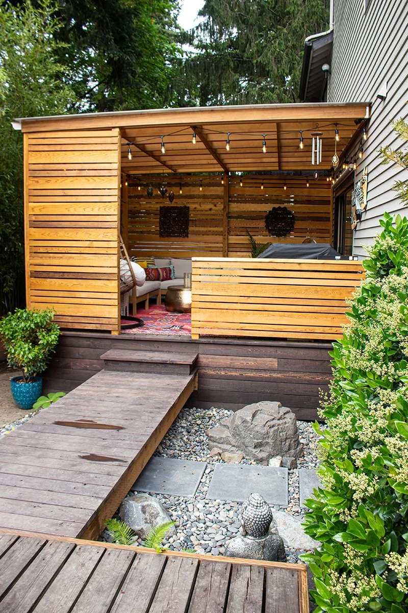 Wooden outdoor patio with seating area, string lights, and decorative elements, adjacent to a house with white siding and surrounded by greenery.