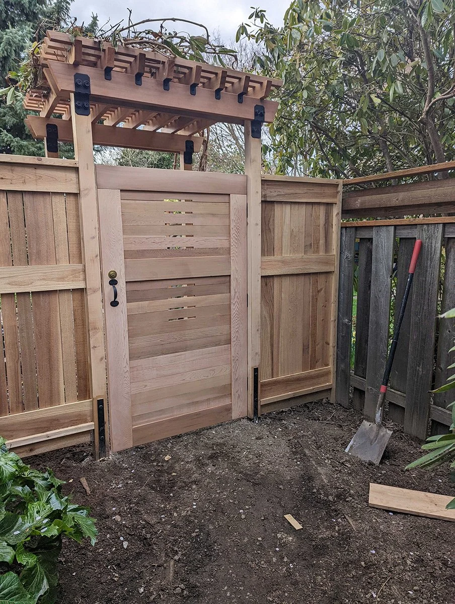 New wooden garden gate with a trellis arch on top, next to a wooden fence, with a shovel leaning against it and some dirt in the yard.