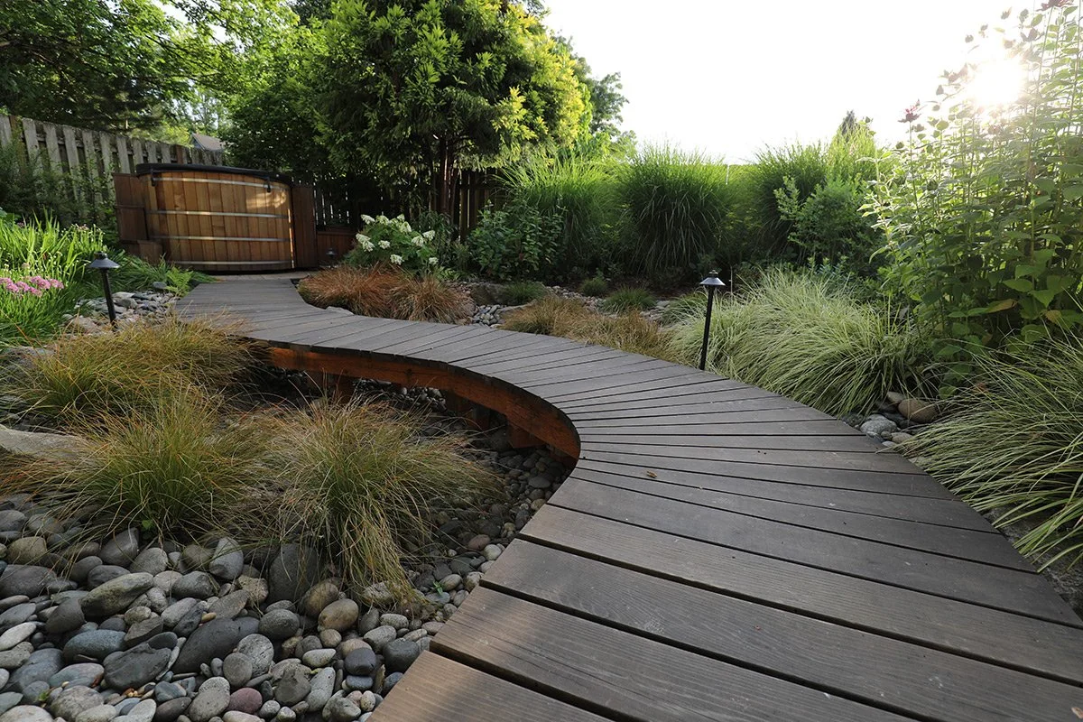 Curved wooden pathway through a landscaped garden with rocks, plants, and small pathway lights, leading to a hot tub in a backyard.