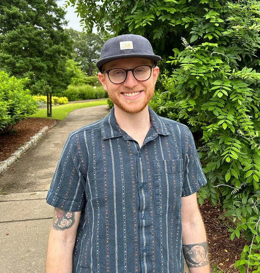 Young man with glasses and a gray cap, smiling outdoors among green trees and bushes.