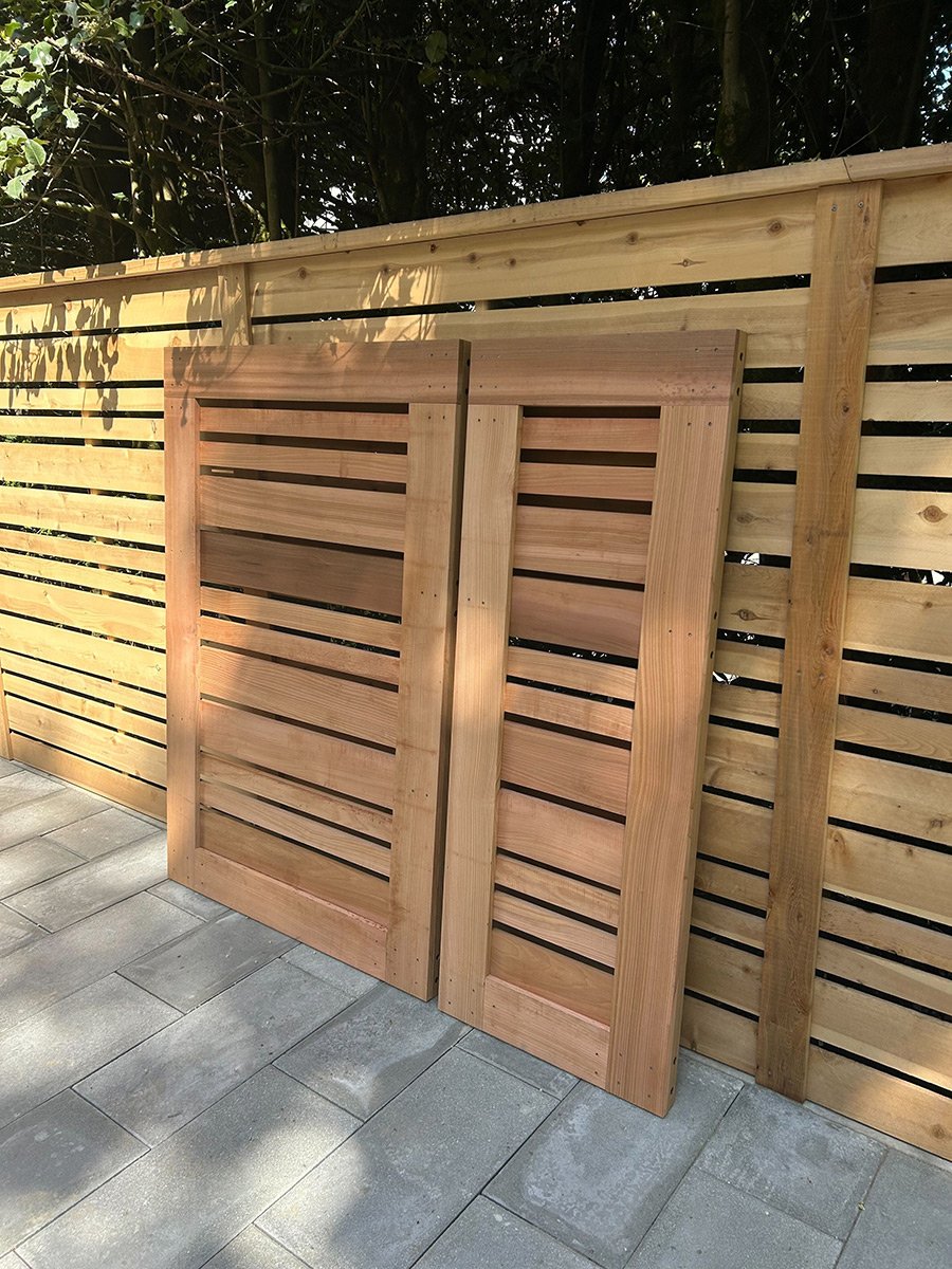Two wooden patio doors leaning against a wooden fence with horizontal slats, outdoors on a paved surface.