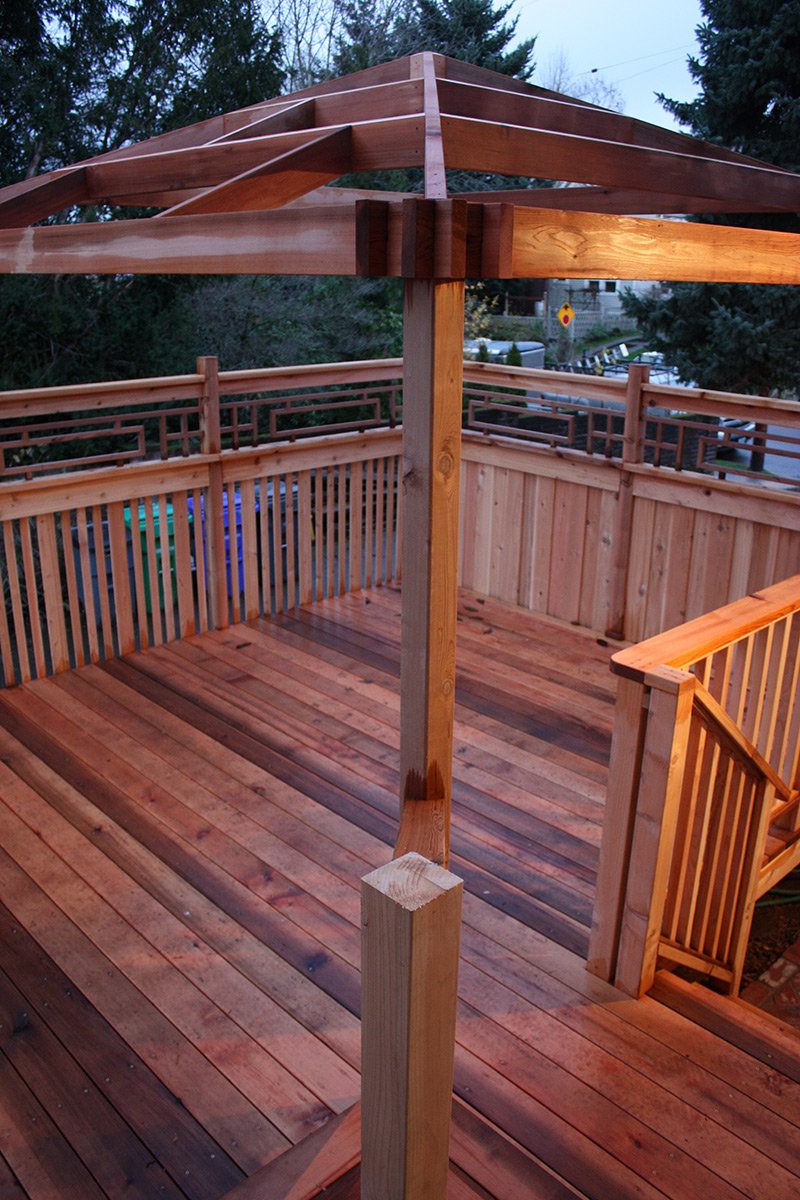 Wooden deck with a partially built gazebo or pergola structure, showing a central support post and an incomplete roof frame, surrounded by a wooden fence and trees in the background.