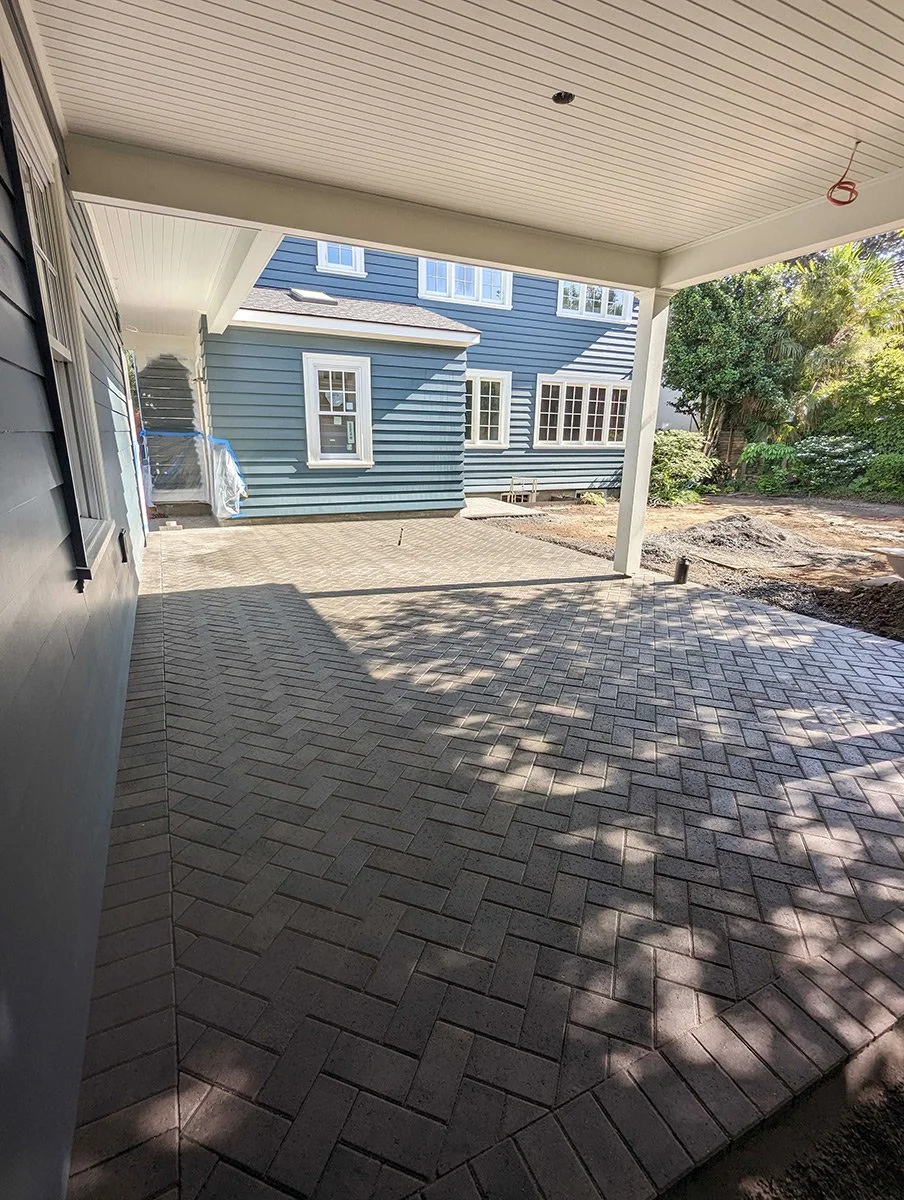 A newly paved backyard patio with interlocking brick pavers, adjacent to a blue house with white trim, and surrounded by trees and greenery.