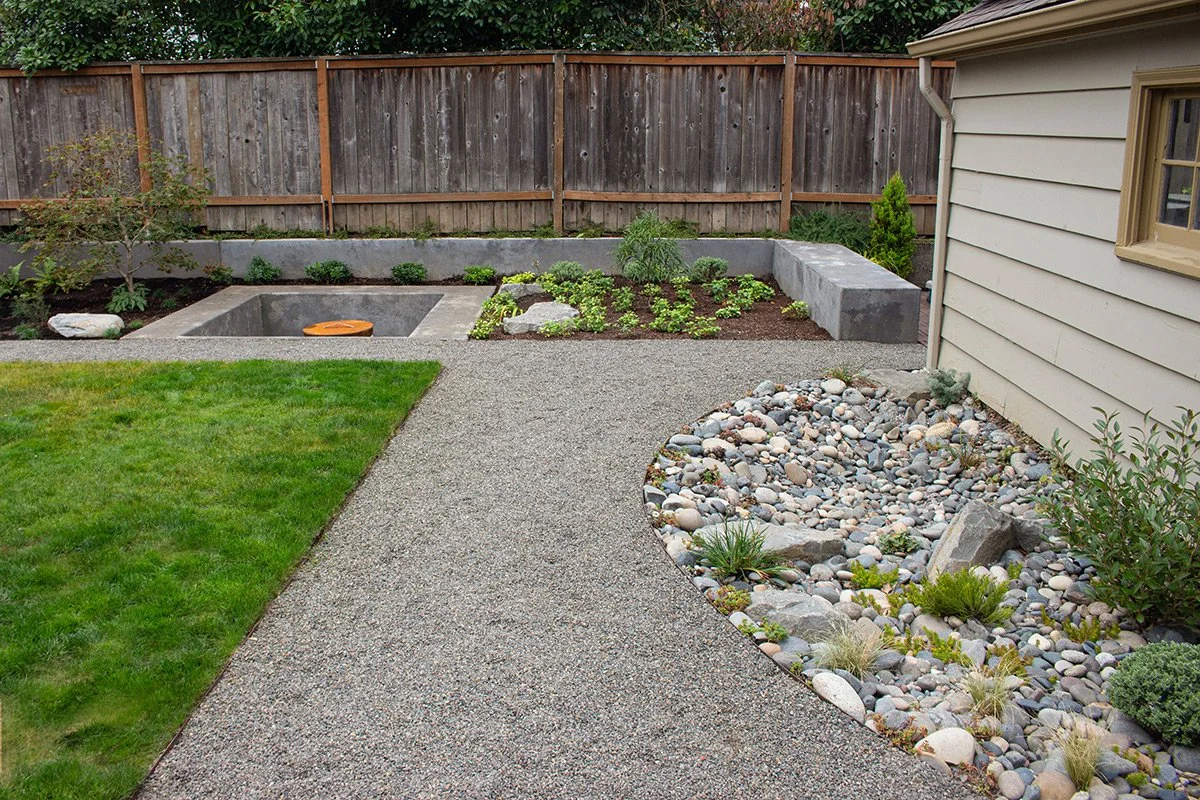 Backyard garden with a gravel pathway, green grass, landscaped rocks, plants, and a wooden fence.