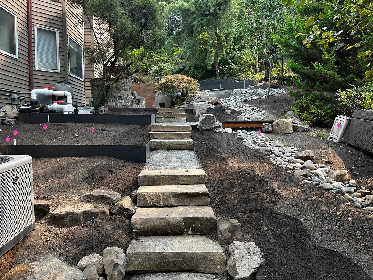 Landscape view of a backyard area under construction with stone steps, black raised garden beds, and a dry riverbed with rocks. Green trees and shrubs surround the scene, with construction materials and pink flags marking areas.