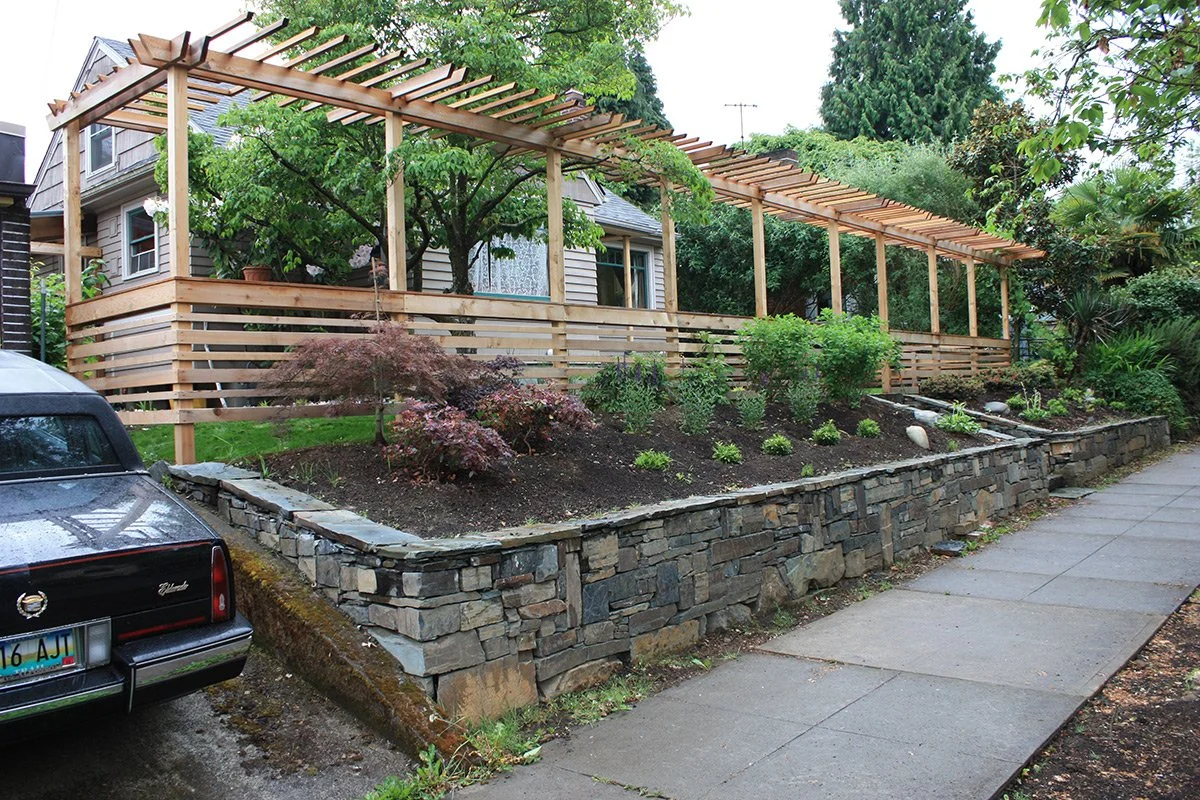 A house with a new wooden deck and pergola extension, adjacent to a stone retaining wall with a garden bed full of plants and shrubs, on a rainy day.