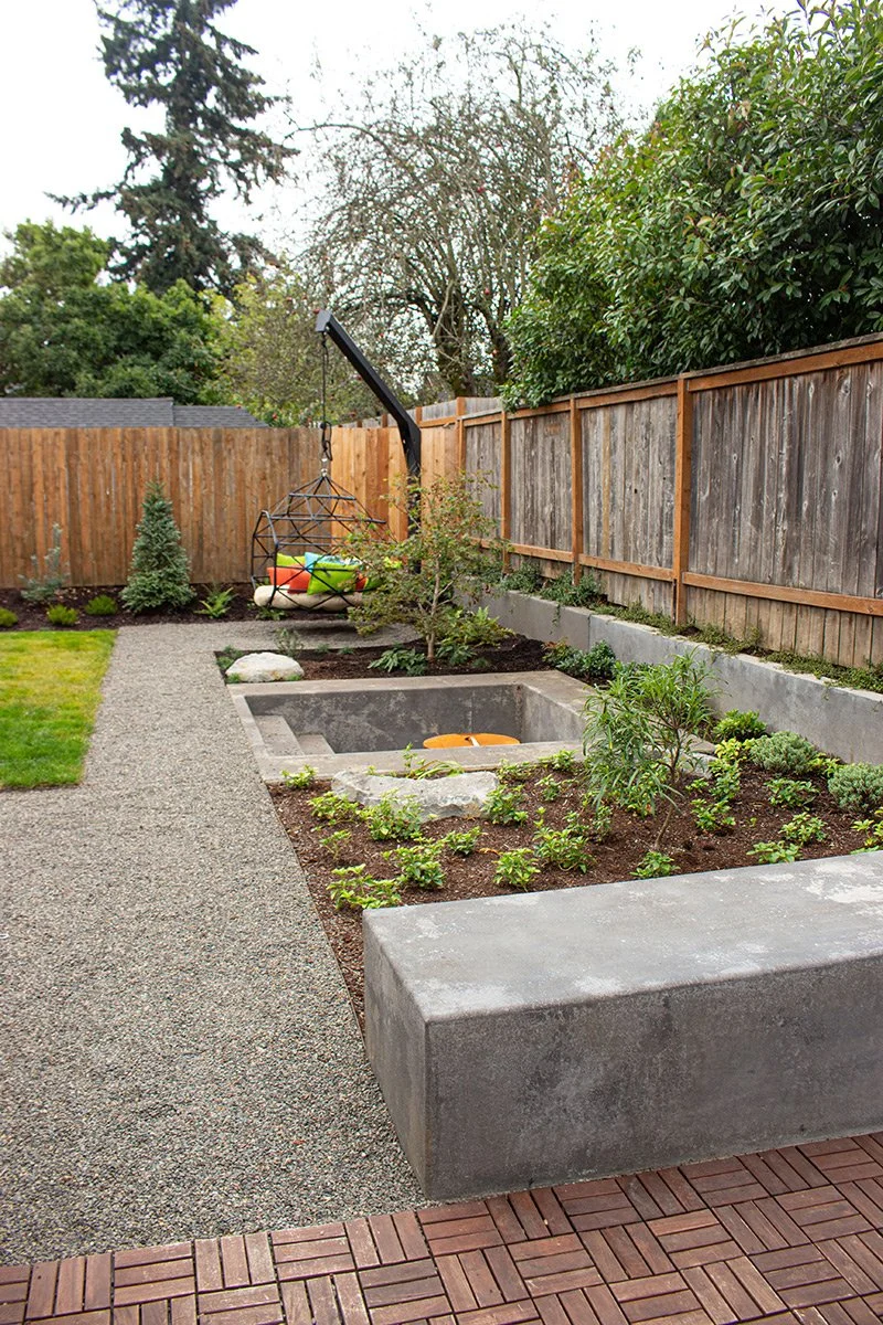 A backyard garden with a gravel pathway, young plants and small trees in mulched beds, a sitting area with a hanging chair and cushions, and a wooden fence surrounding the space.