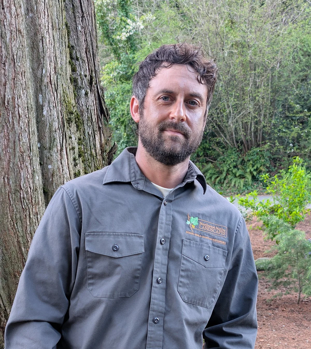 A man with a beard and short brown hair, wearing a gray button-up shirt with a logo, standing outdoors near a large tree and green shrubbery in a natural setting.