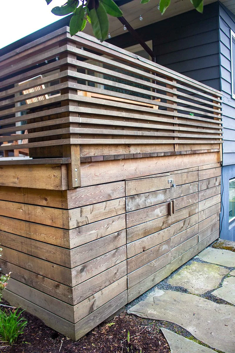 Wooden structure with horizontal slats on top and vertically arranged wooden planks below, located outside a house.